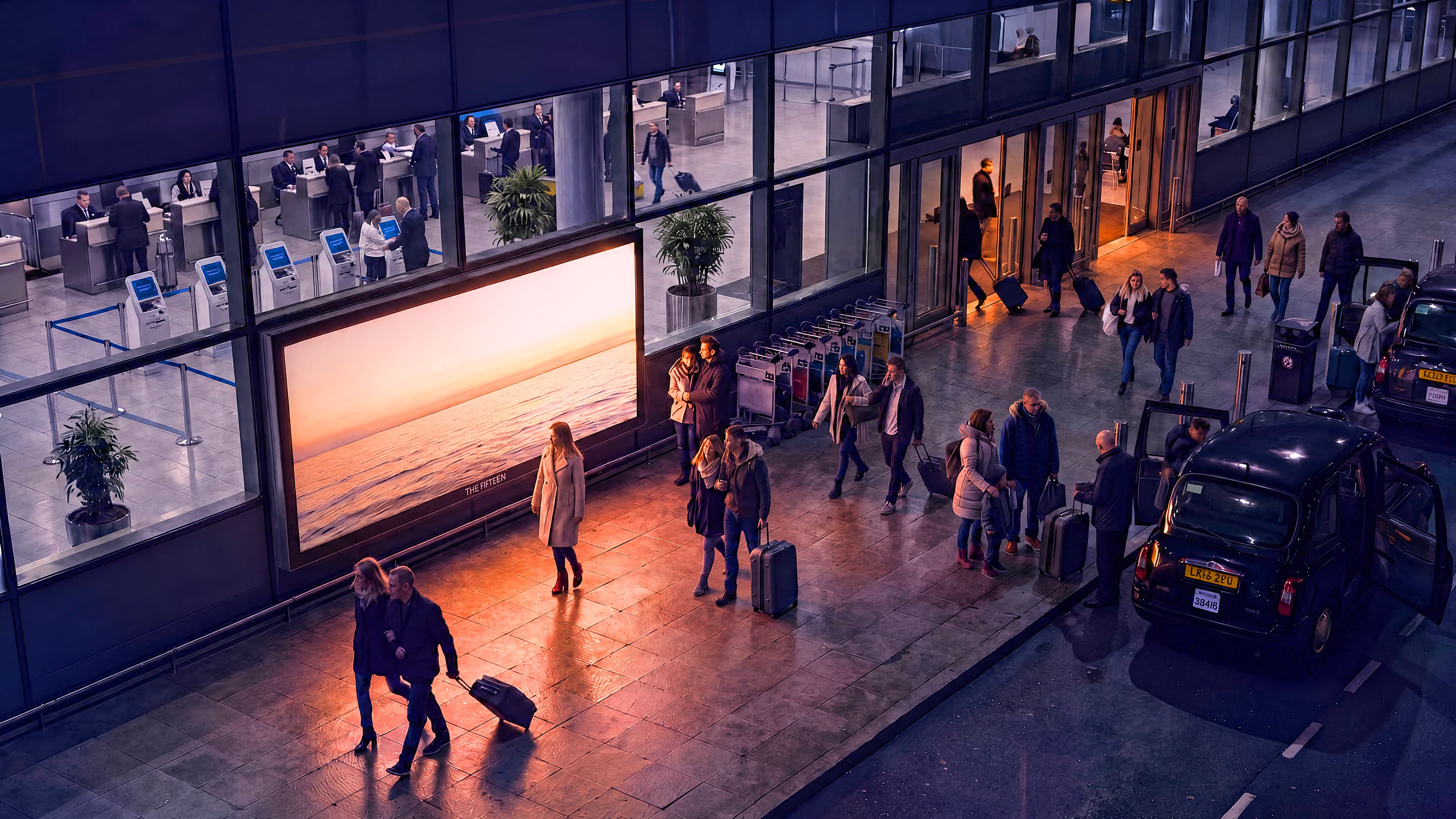 Travellers cross a rain-slicked terminal kerb at night while a large illuminated seascape advertisement glows unwatched beside the doors; a black cab loads at the rank.