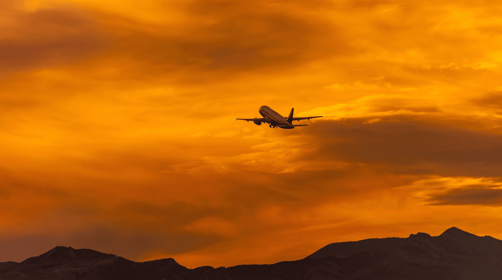 A plane climbing into an orange sunset sky above dark mountains